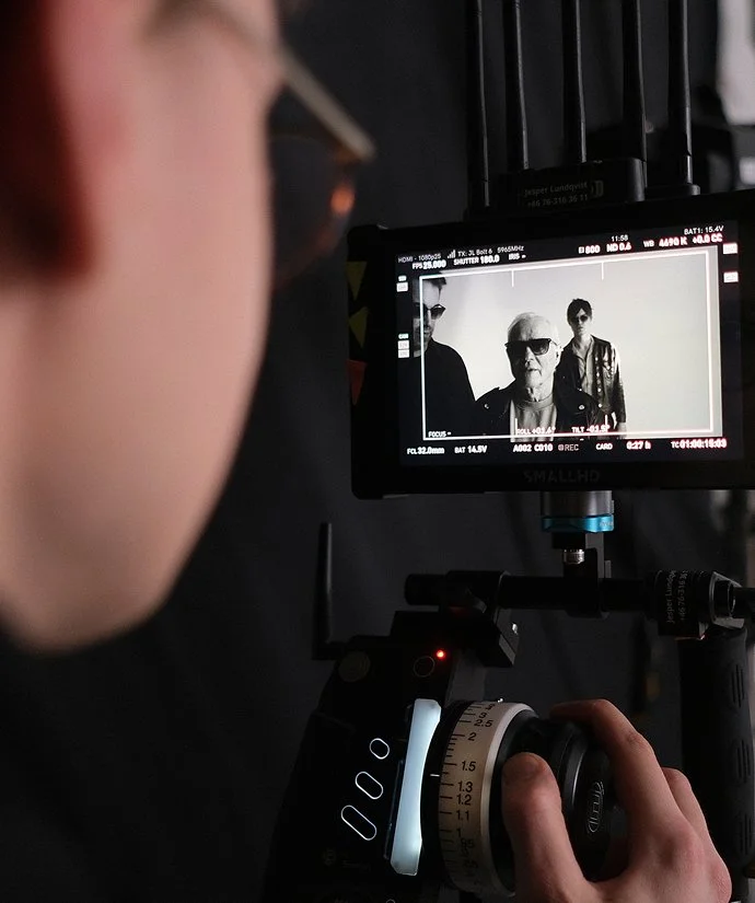 Person som filmar tre personer i svartvitt på en skärm, där en av personerna är äldre och bär solglasögon.