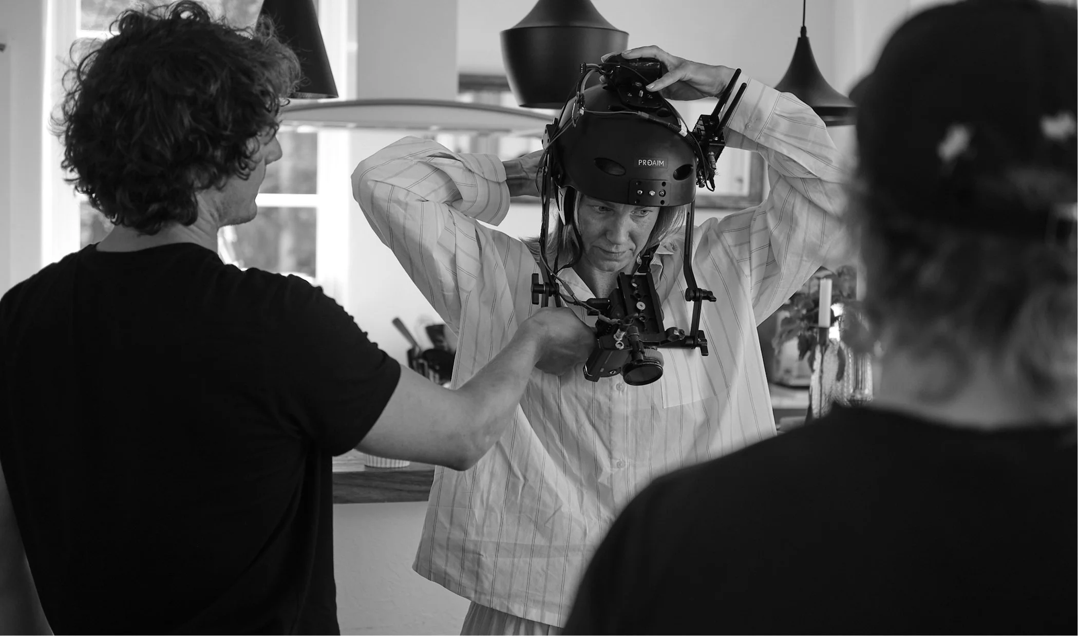 En person justerar en helikopterhjälm med en kamerautrustning medan två andra personer assisterar, i ett inomhusmiljö, svartvitt fotografi.