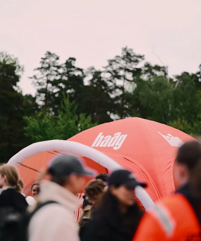 En grupp människor samlade runt en orange tältliknande struktur med texten 'haag' på, utomhus med träd i bakgrunden.