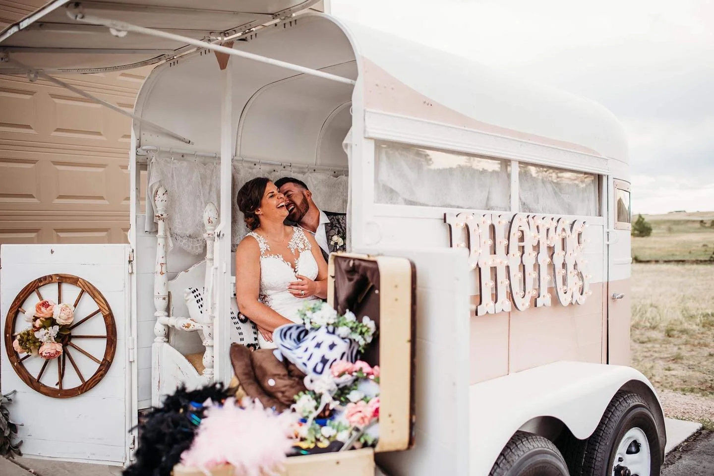A bride and groom laughing together inside a vintage white trailer decorated with flowers and a sign that says 'LOVE'