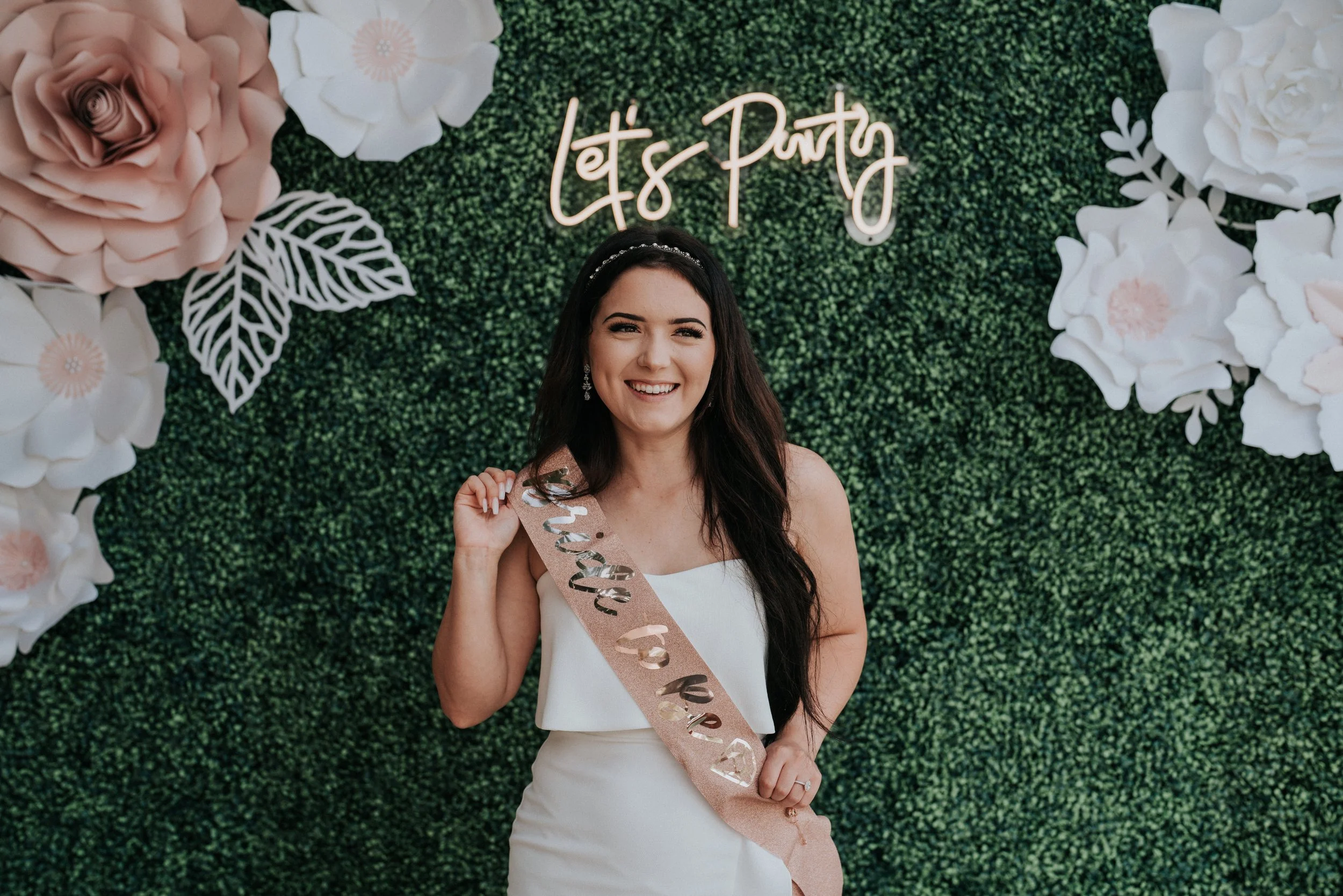 Woman wearing a 'bride to be' sash at a bridal shower, standing in front of a green floral wall with large paper flowers and a neon sign that says 'Let's Party'.