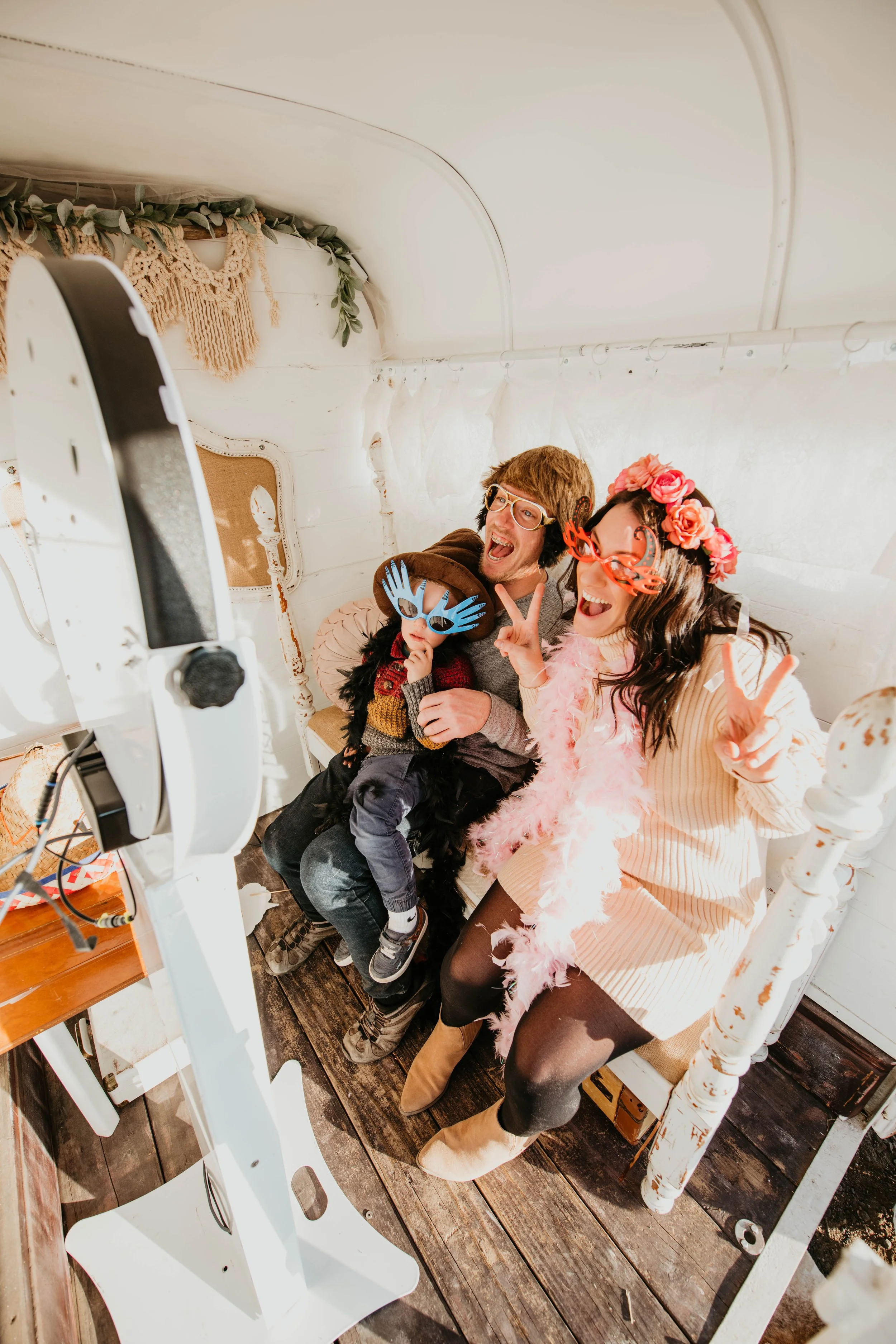 Three people in costume posing for a photo in a photo booth, wearing funny glasses, feather boas, and floral headpieces, with a camera on a stand in front of them.