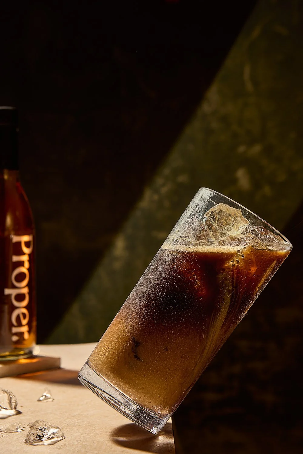 Leaning glass of iced coffee with a bottle of Proper Syrup in the background
