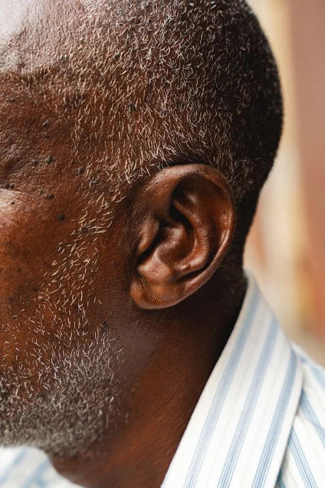 Close-up of a man's side head showing short, graying hair and a brown ear, with part of a striped shirt visible.