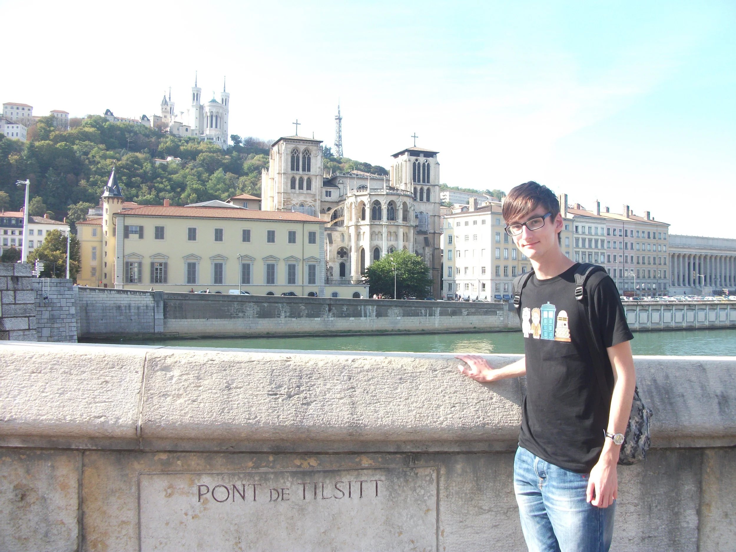 Luke standing at the Pont de Tilsitt, an old stone bridge over the river Saône in Lyon, France.  The sun is shining over Vieux-Lyon architecture in the background, including the Fourvière basilica atop a wooded hill.