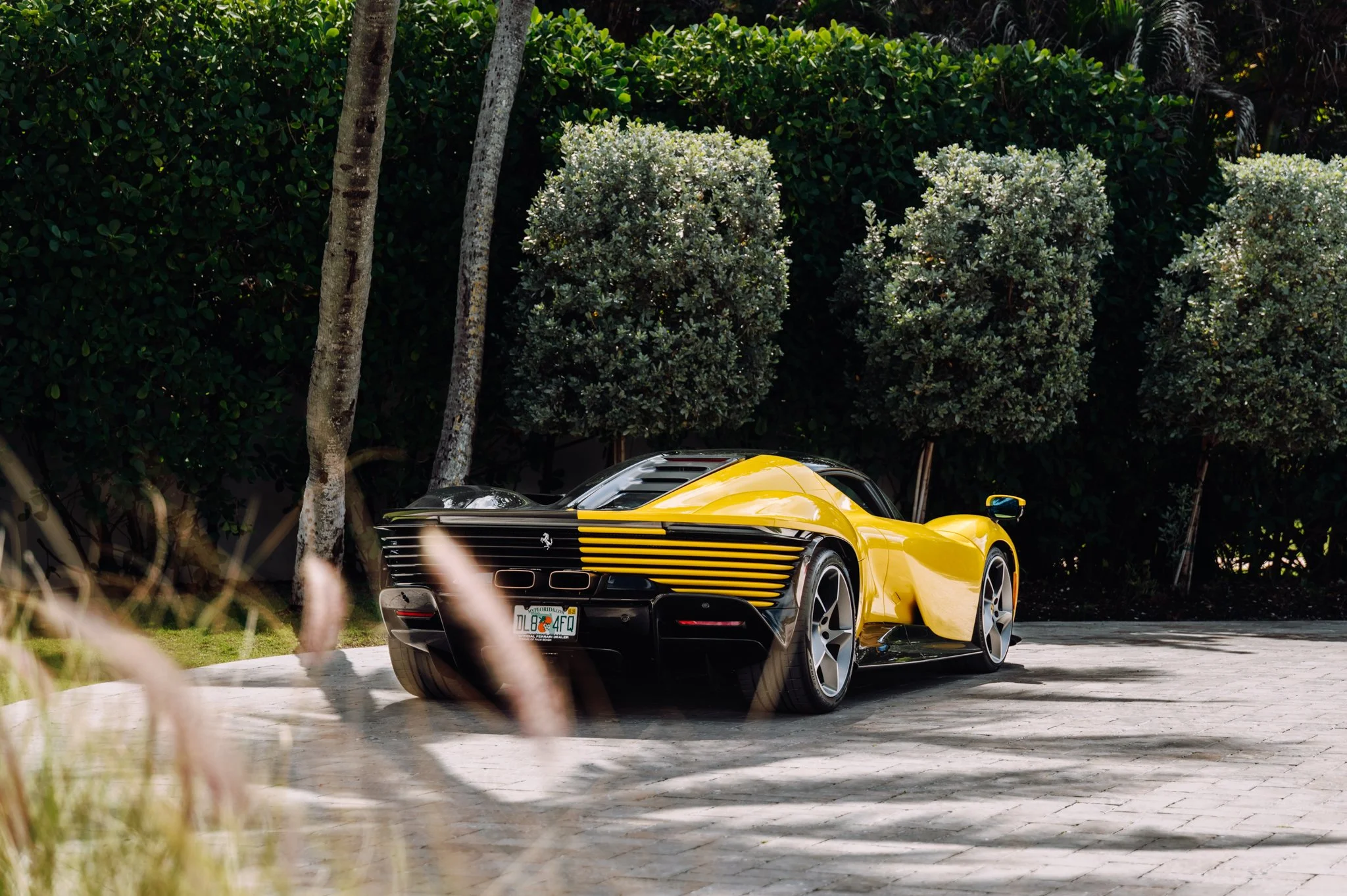 A yellow sports car parked on a driveway with tall trees and green bushes in the background.