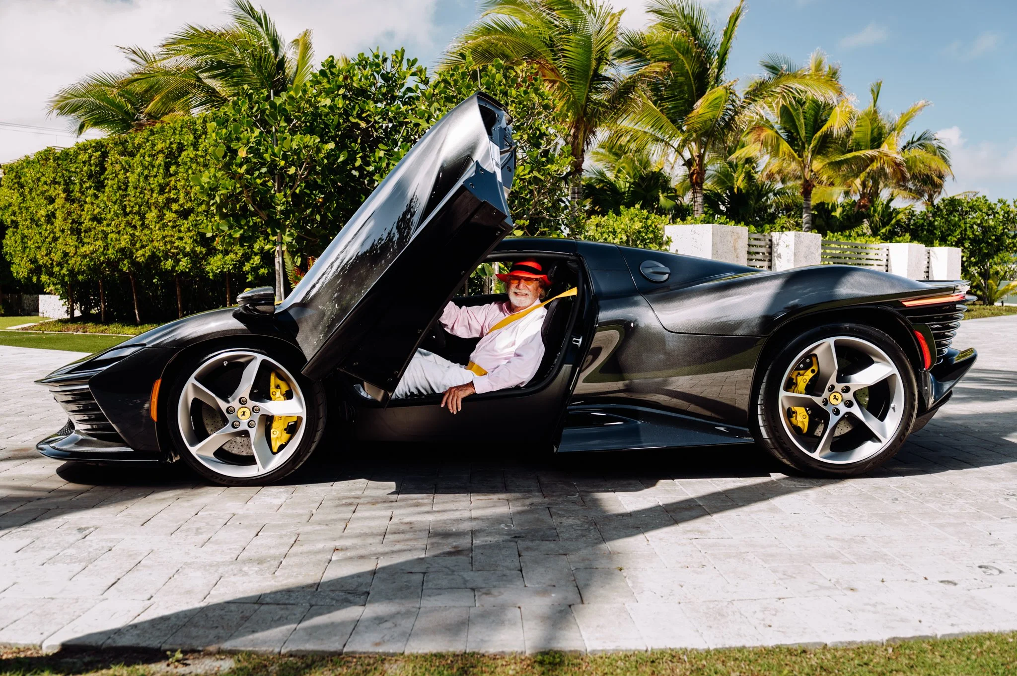 A man with a white beard and wearing sunglasses, a pink shirt, and a red hat, sitting in a black luxury sports car with gullwing doors open, parked outdoors with palm trees and greenery in the background.