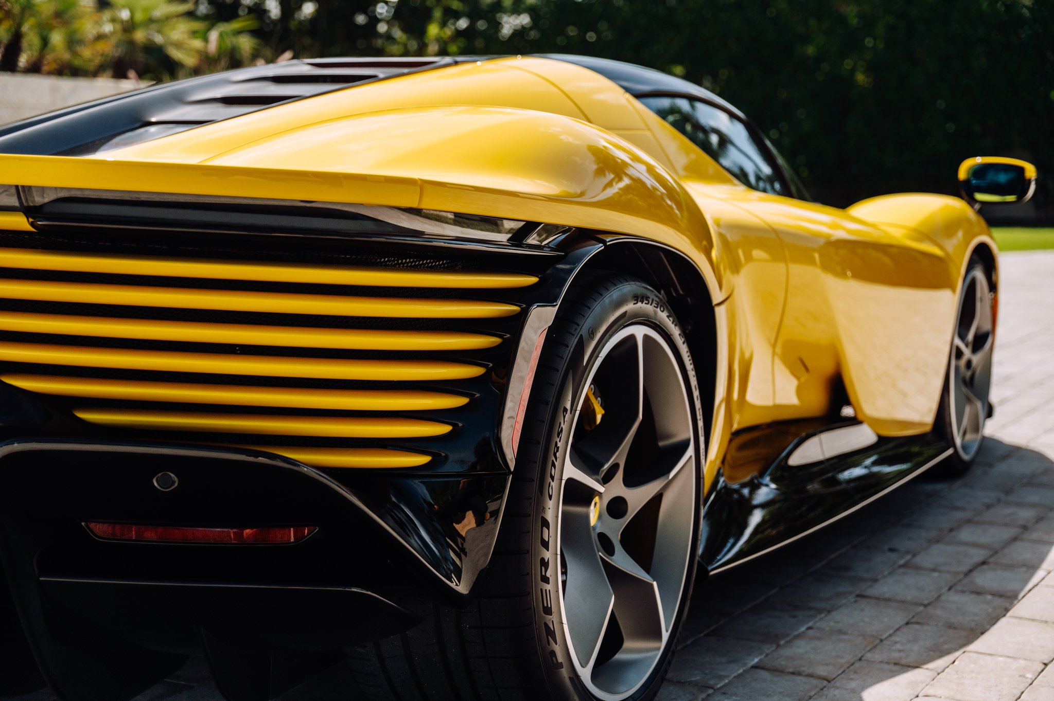 Close-up of a yellow sports car showing the front grille, headlight, and part of the wheel, parked on a paved surface with greenery in the background.