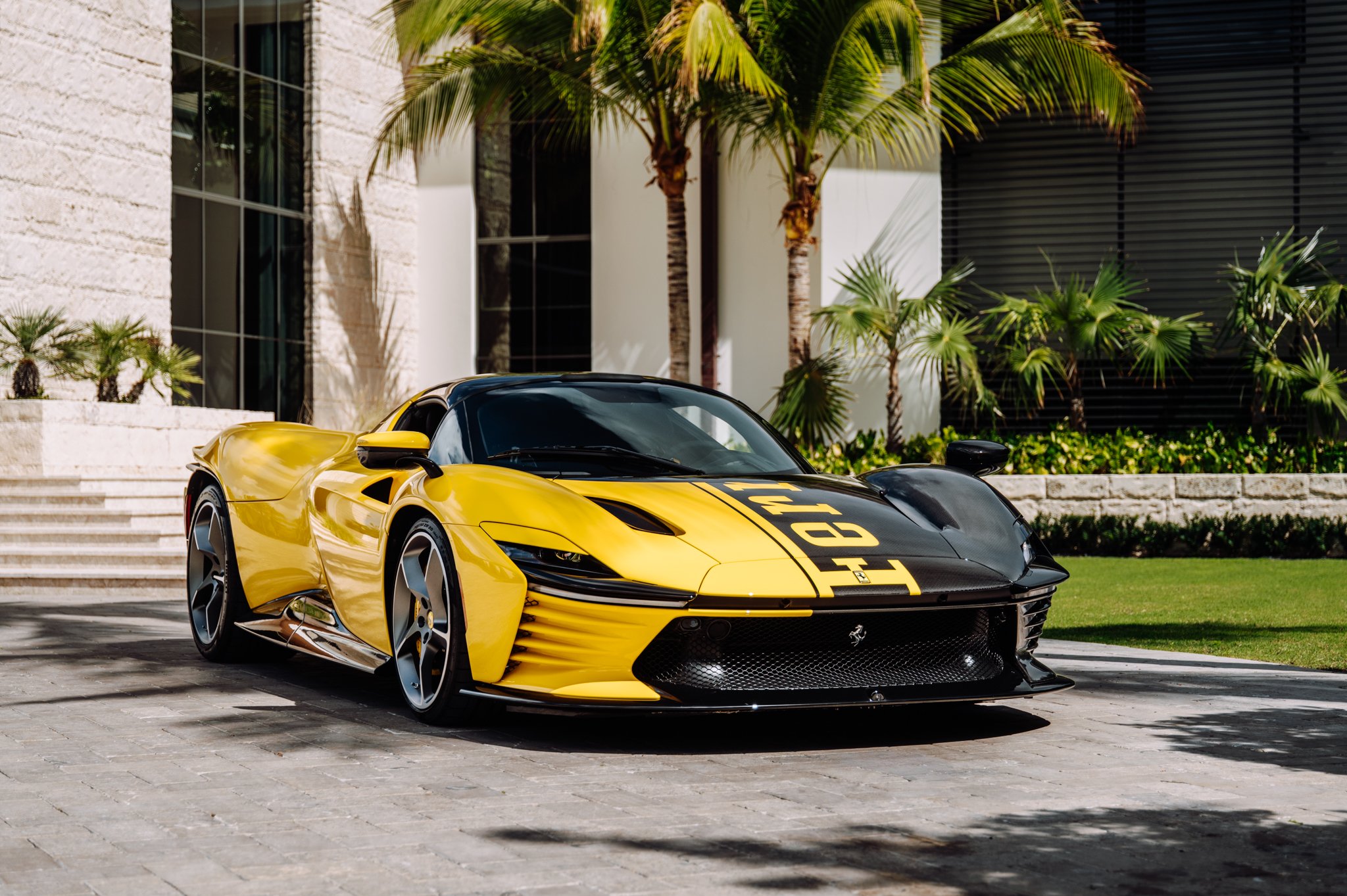 A yellow and black Ferrari sports car parked outside a modern building with large windows, palm trees, and lush greenery in the background.