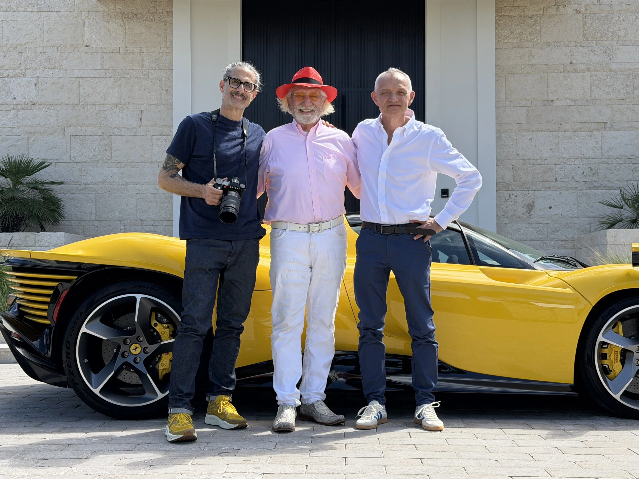 Three men standing in front of a yellow sports car, posing with arms around each other, smiling, outdoors in front of a modern building.