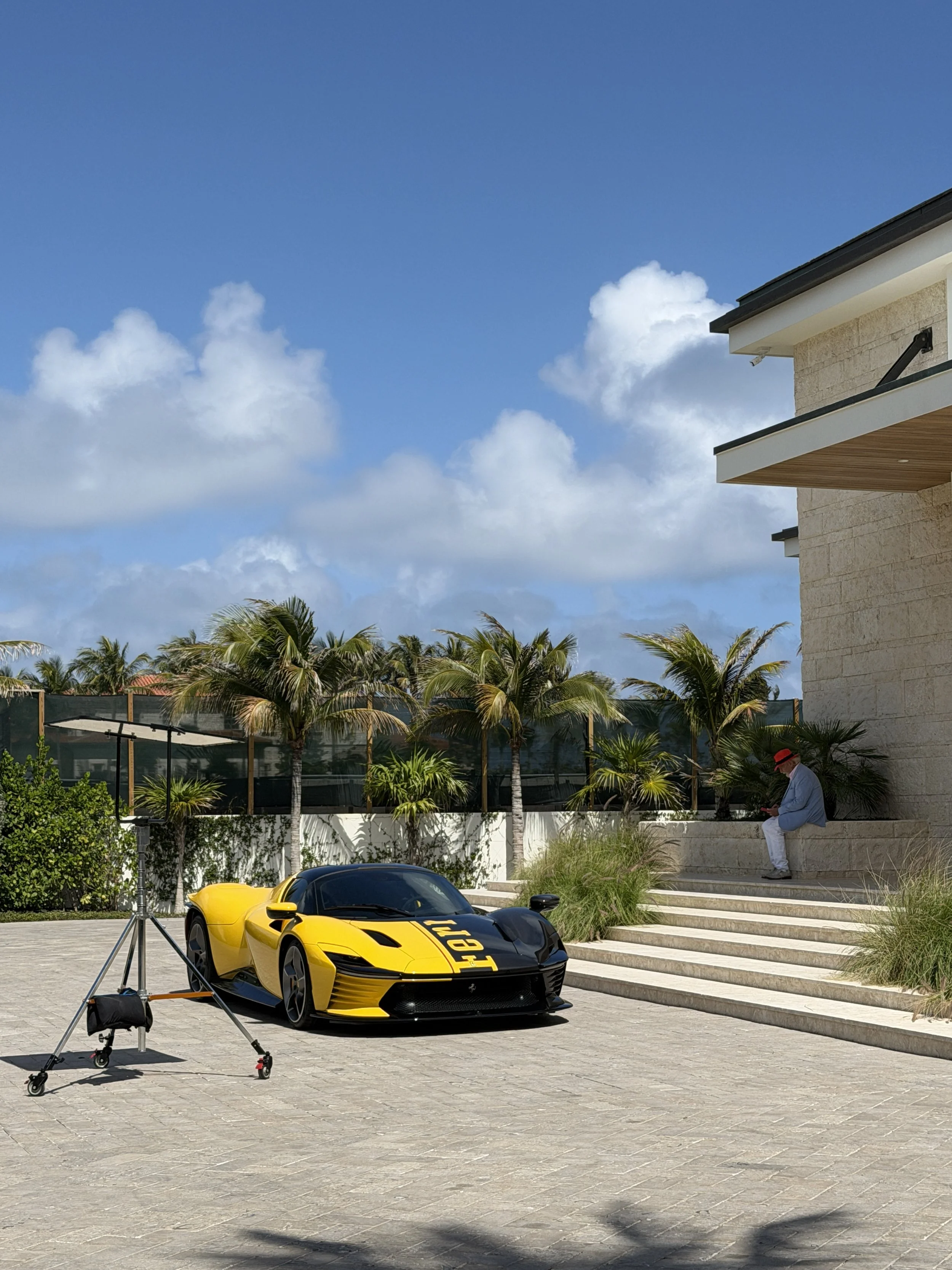 A yellow Lamborghini sports car parked on a paved driveway, with palm trees in the background and a modern house with a balcony on the right. A man in a white suit and red hat is sitting on steps, looking at his phone, and a photographer's tripod is 