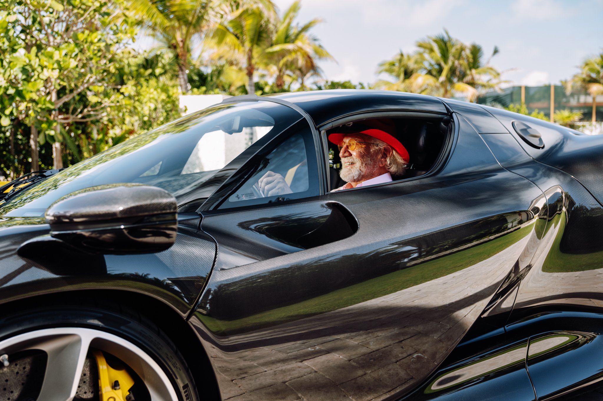 A man with a beard and sunglasses sitting in a black sports car, wearing a red hat, white shirt, and pink tie, with a background of palm trees and a blue sky.
