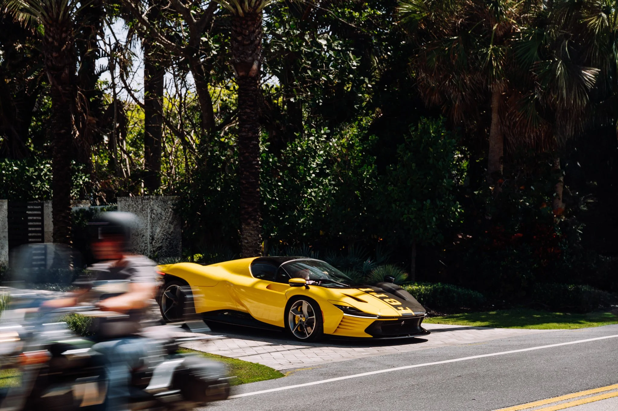 A yellow luxury sports car parked on the side of a street with tall trees and green foliage in the background, while a motorcycle and a car pass by quickly in the foreground.