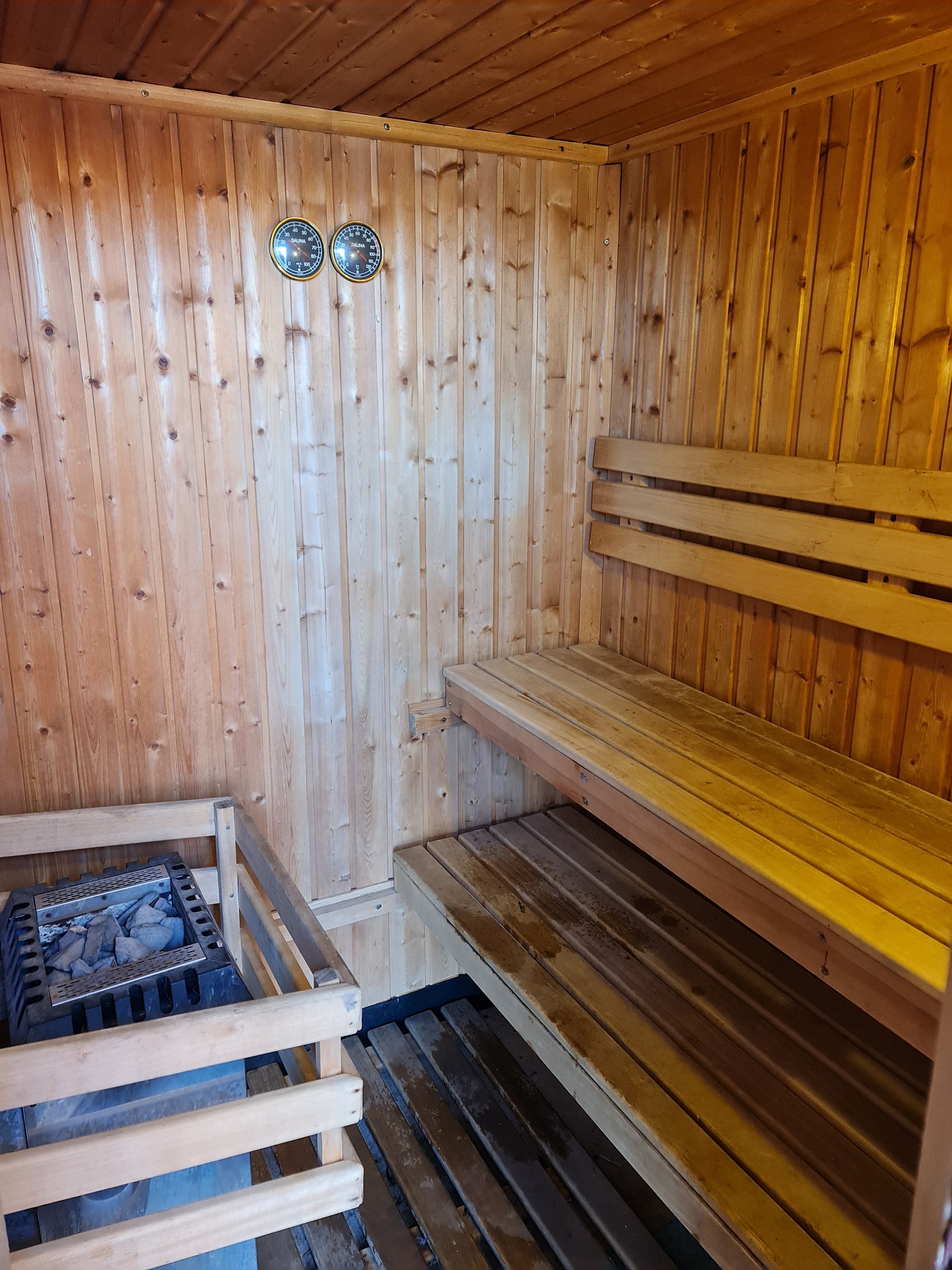 Inside a traditional wooden sauna with wooden benches, a heater with stones, and a thermometer and hygrometer on the wall.