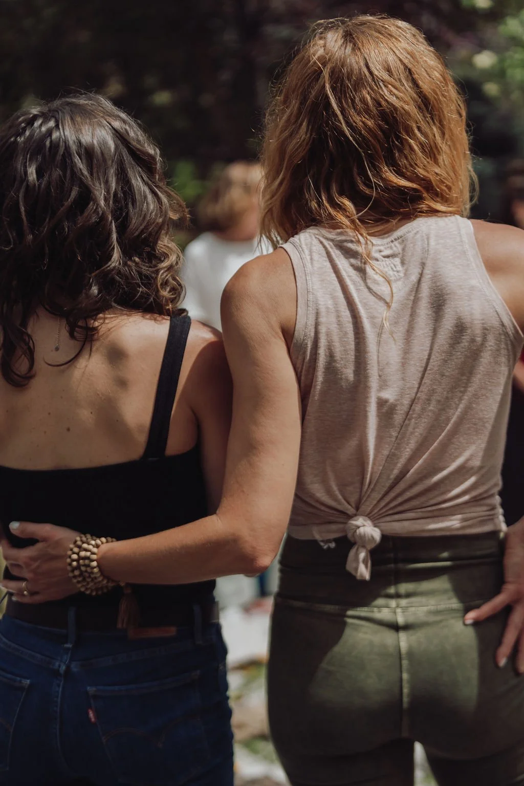 Two women standing close together outdoors, one with dark wavy hair wearing a black tank top and denim jeans, the other with reddish hair wearing a sleeveless beige top and dark pants.
