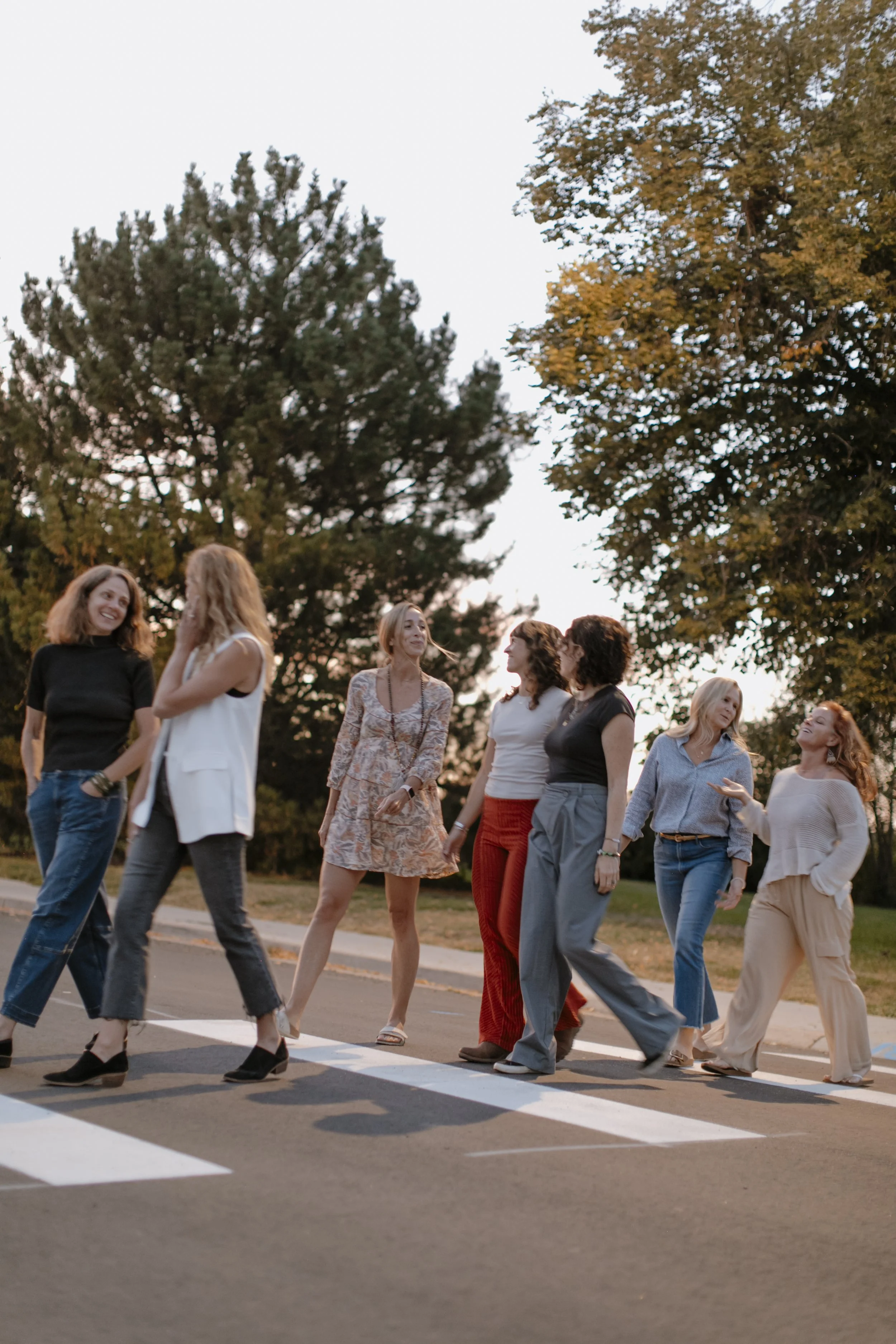 A group of women walking and talking in a park or outdoor area during sunset, surrounded by trees with some autumn foliage.