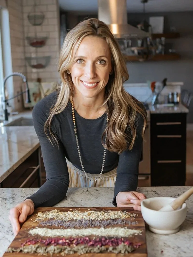 A woman with long, wavy blonde hair and a black shirt smiling while presenting a wooden platter of layered desserts in a kitchen.