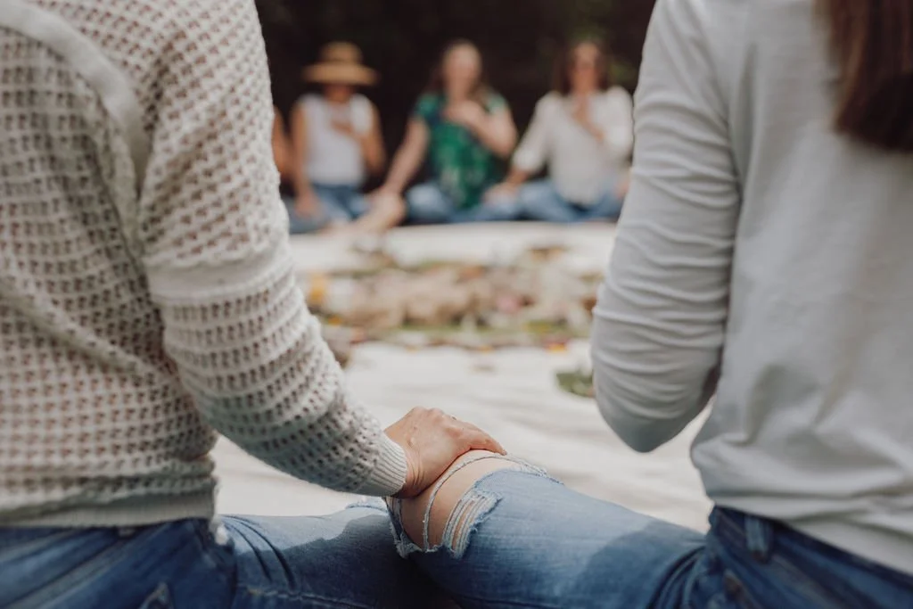 People sitting outdoors on a blanket, having a picnic, with a group in the background.