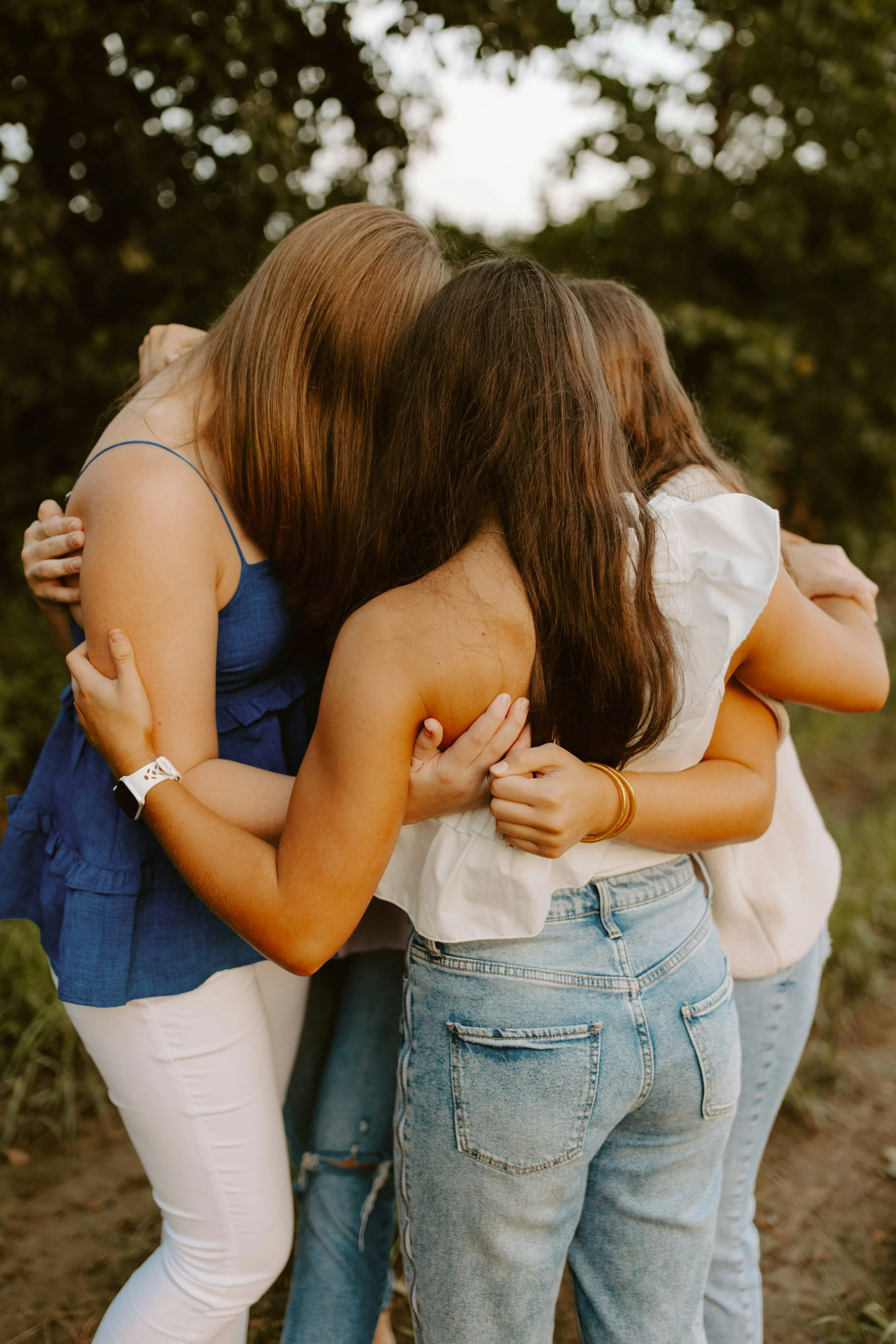 A group of women wearing casual clothing hugging with their eyes closed outdoors on a cloudy day.