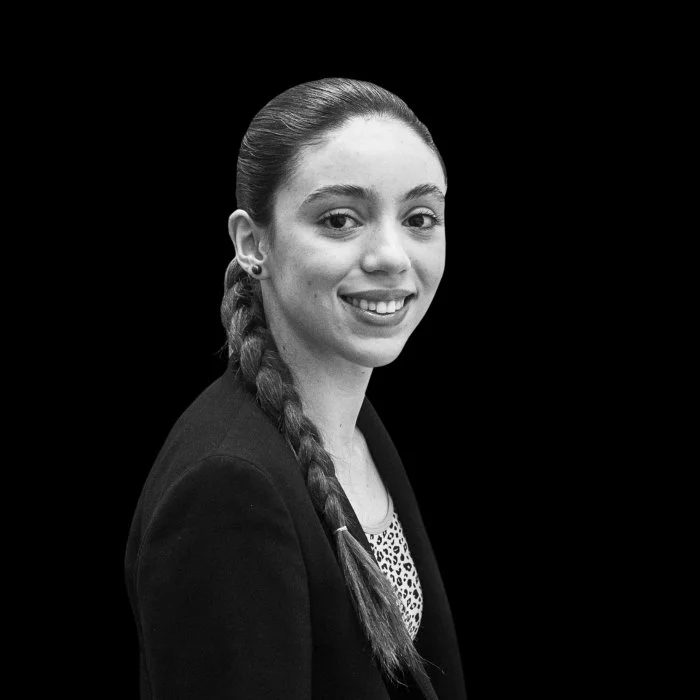 A woman with short curly hair wearing glasses, hoop earrings, and layered necklaces smiling in an indoor setting.