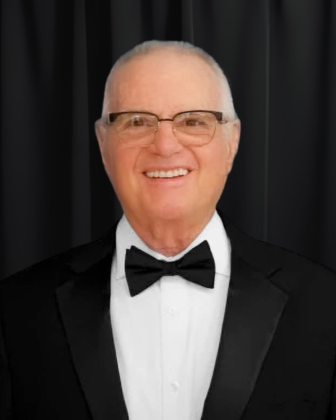 A smiling older man wearing glasses, a tuxedo, white dress shirt, and black bow tie, photographed against a black background.