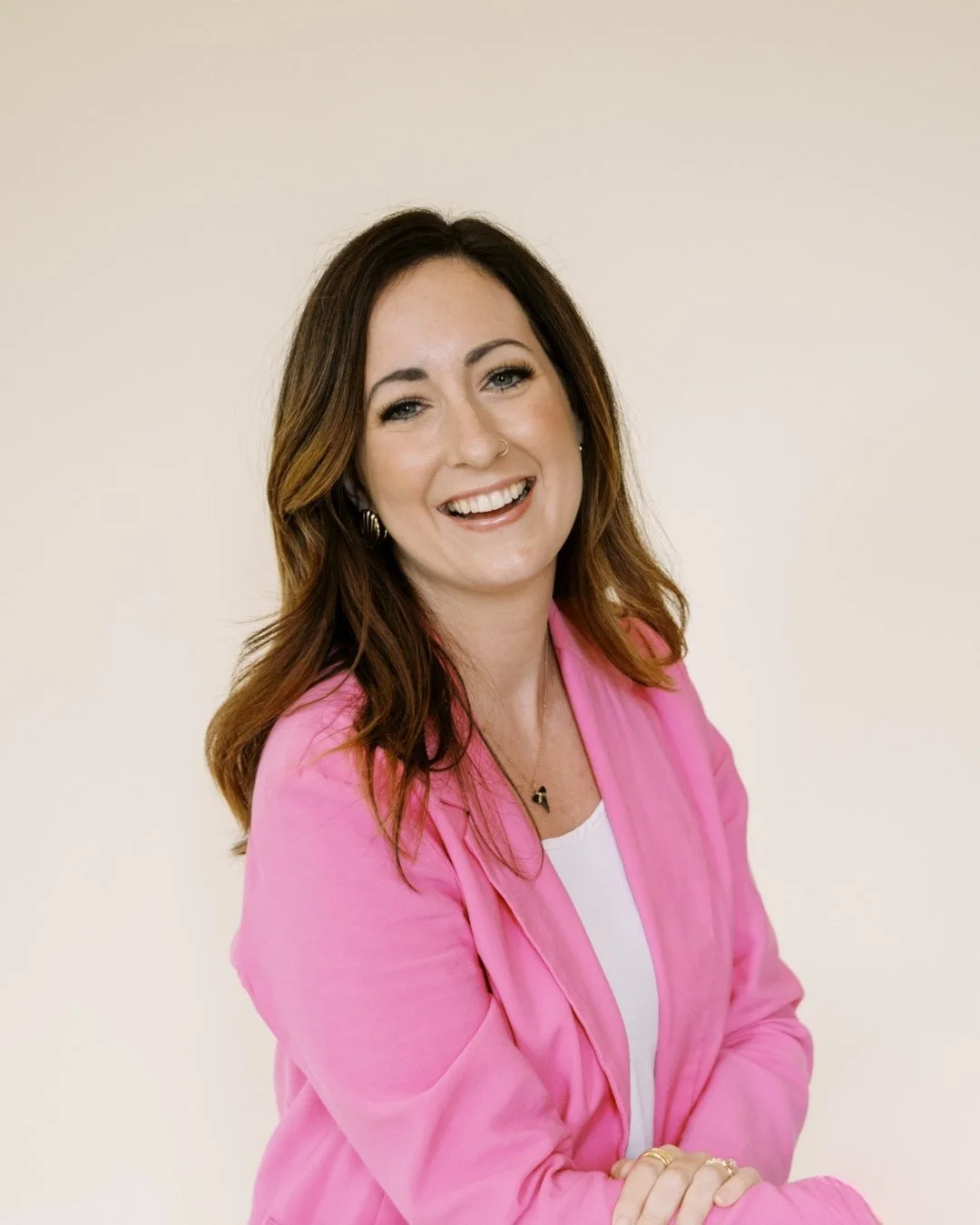 A young woman with long brownish hair smiling, wearing natural makeup, a white top, and a bright pink blazer.