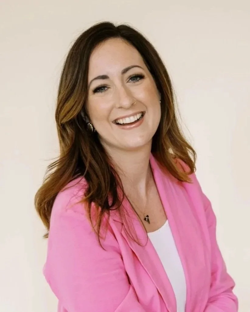 Close-up of a young woman with long brownish hair smiling, wearing natural makeup, a white top, and a bright pink blazer.