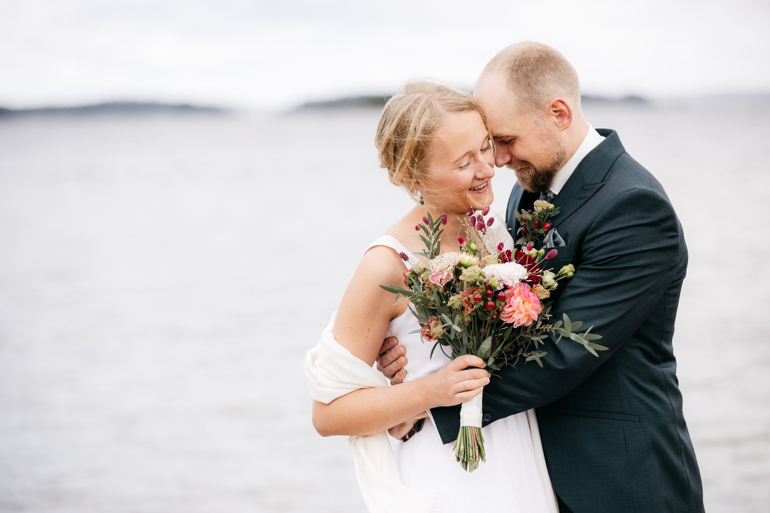 Ett brudpar som håller varandra nära vid havet, båda ler och är nära varandra, bruden håller en bukett med rosa och vita blommor.