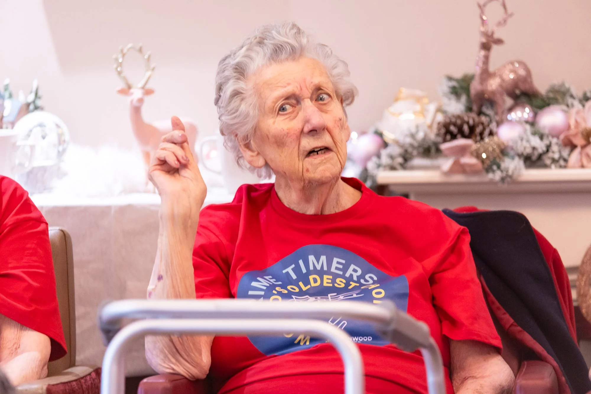 Elderly woman in red shirt gesturing with hand, seated near festive decorations, and using a walker.