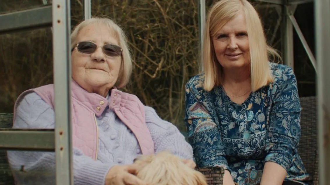 Two women sitting with a small dog, one wearing sunglasses and a pink vest, the other in a blue patterned dress.