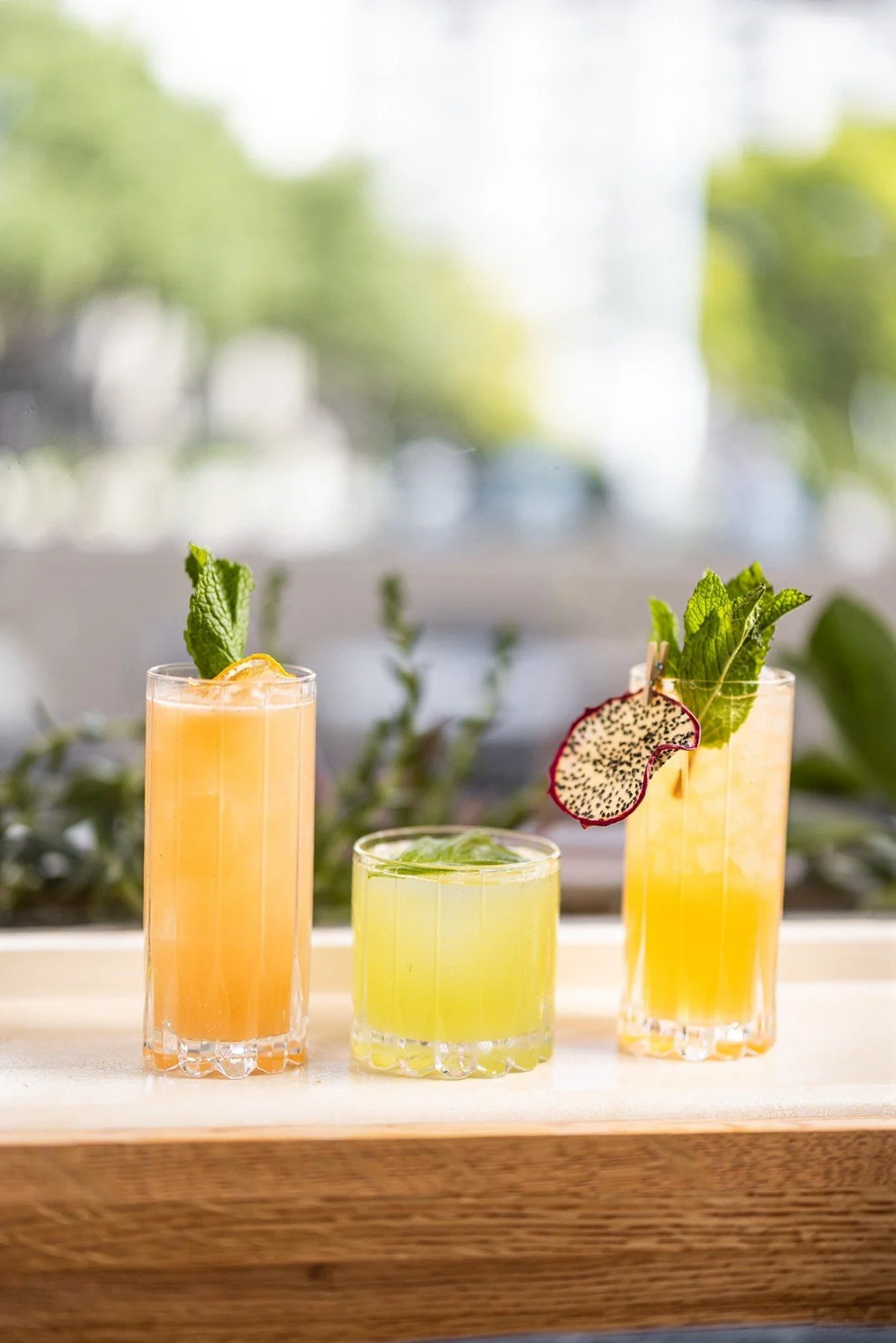 Three cocktails on a wooden table, each garnished with mint. One has orange juice, one is green, and another is garnished with a slice of dragon fruit.