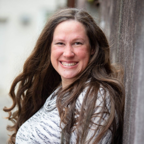 Leslie Davis, Brunette in white and grey striped shirt leaning against a grey or wood wall.