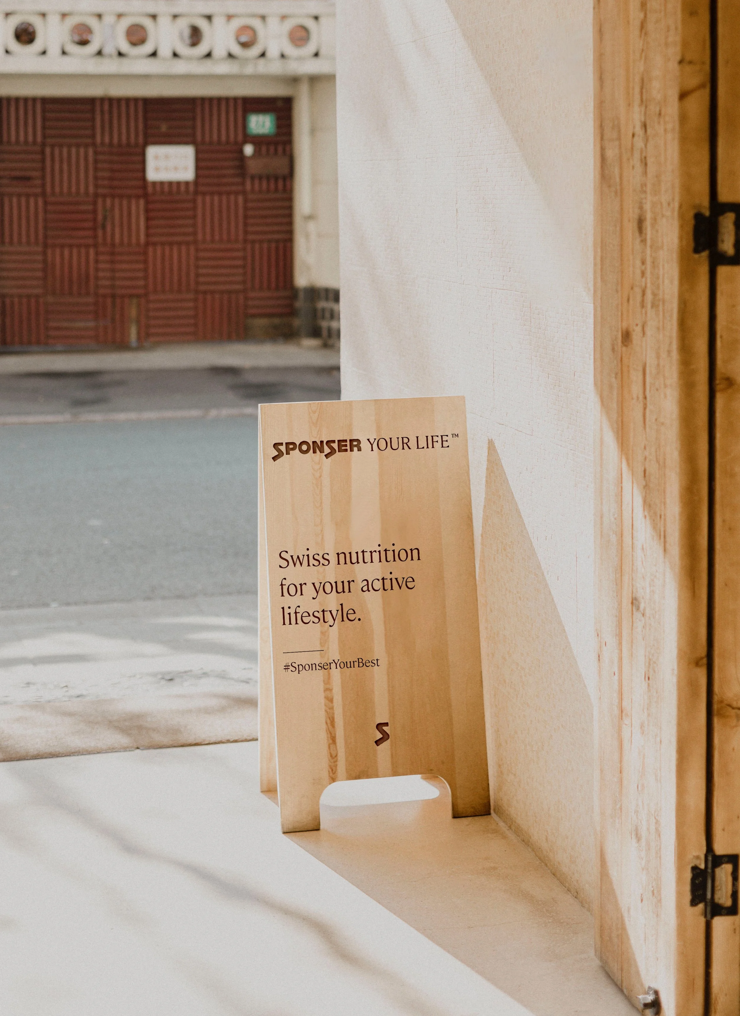 Ein Werbeschild mit dem Text: 'Sponser Your Life. Swiss nutrition for your active lifestyle. #SponserYourBest', steht in einem Raum neben einer Wand und einem Fenster, durch das eine Straße im Freien sichtbar ist.