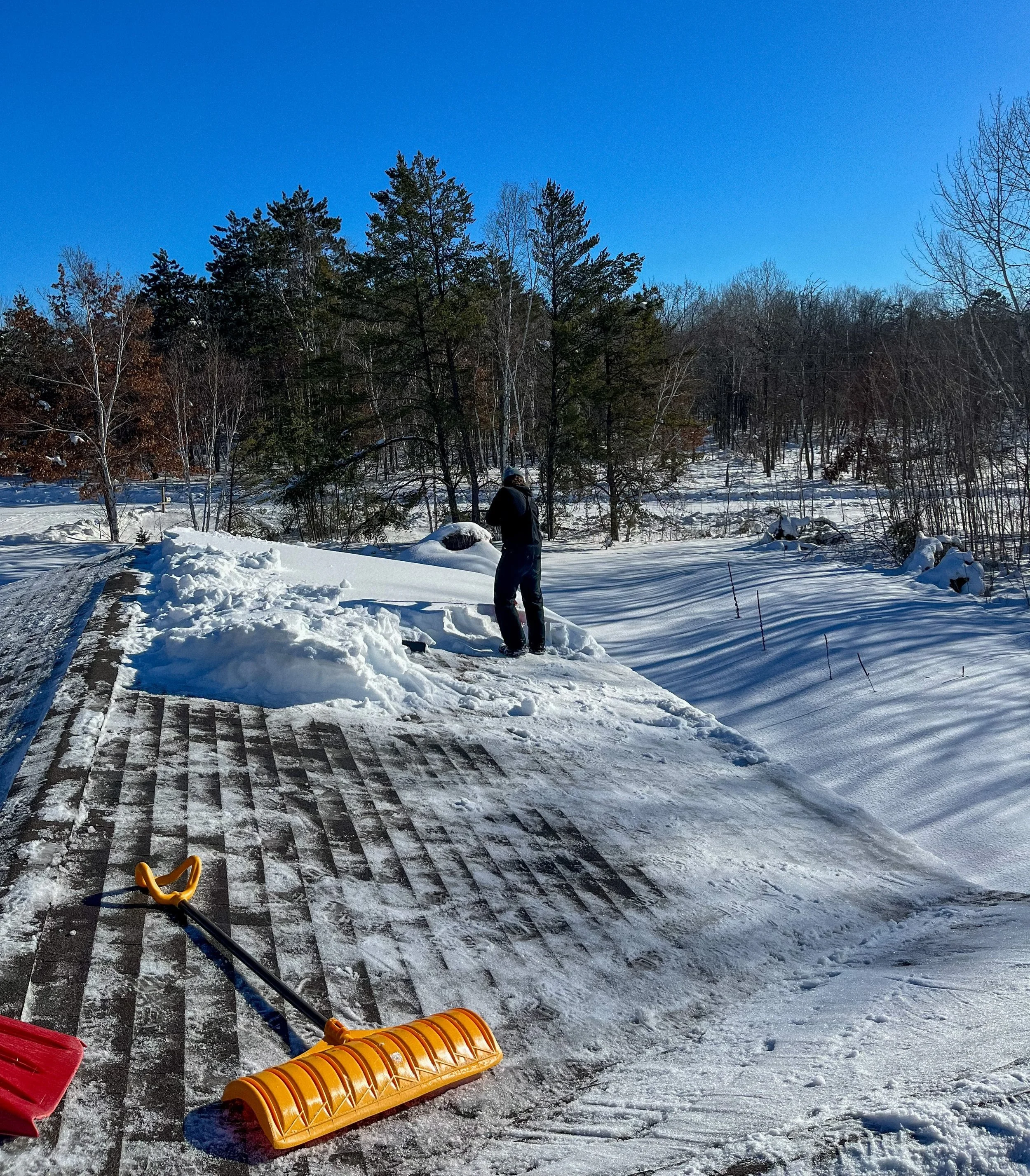 A person shoveling snow off a wooden deck in winter, with snow-covered trees and a clear blue sky in the background.
