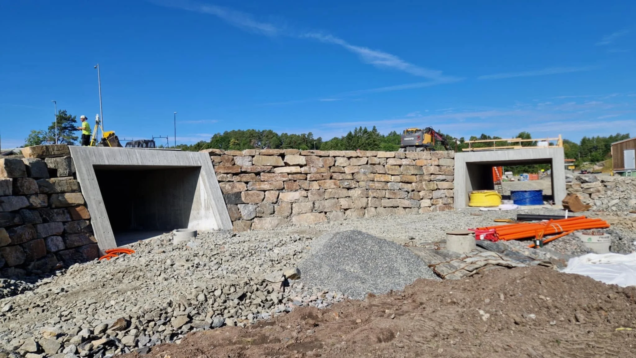 Et byggeområde med en lav mur av stein og betongstrukturer, byggeutstyr og materiell spredt rundt, under en klar blå himmel.