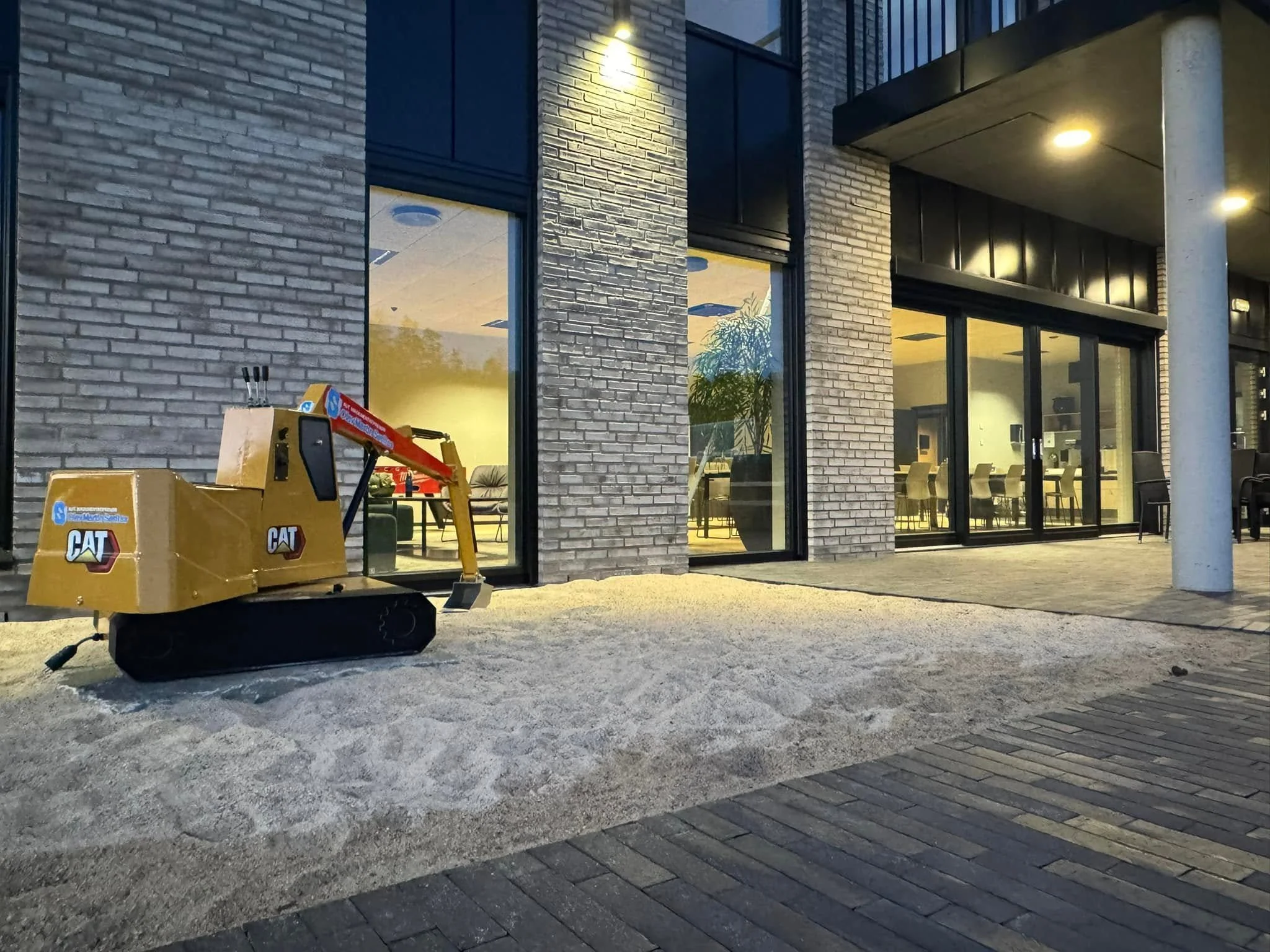Bygning med store glassvinduer og utendørs belysning, fjelltog på bakken, og stillestående kjøretøy i en oppkjørsel.