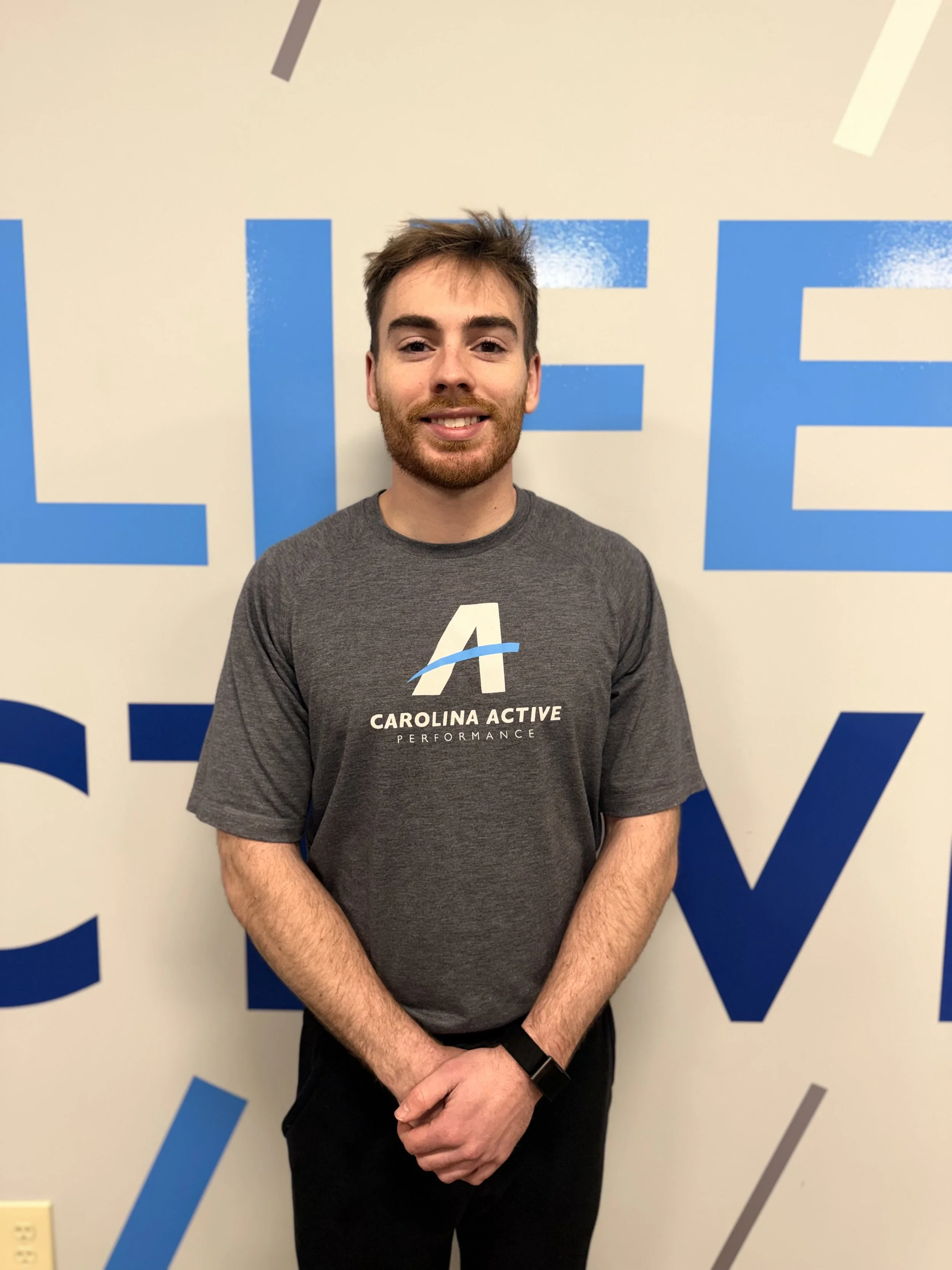 A young man with brown hair, a beard, and a mustache, wearing a gray T-shirt with 'Carolina Active Performance' and an 'A' logo, standing in front of a wall with large blue and gray letters.