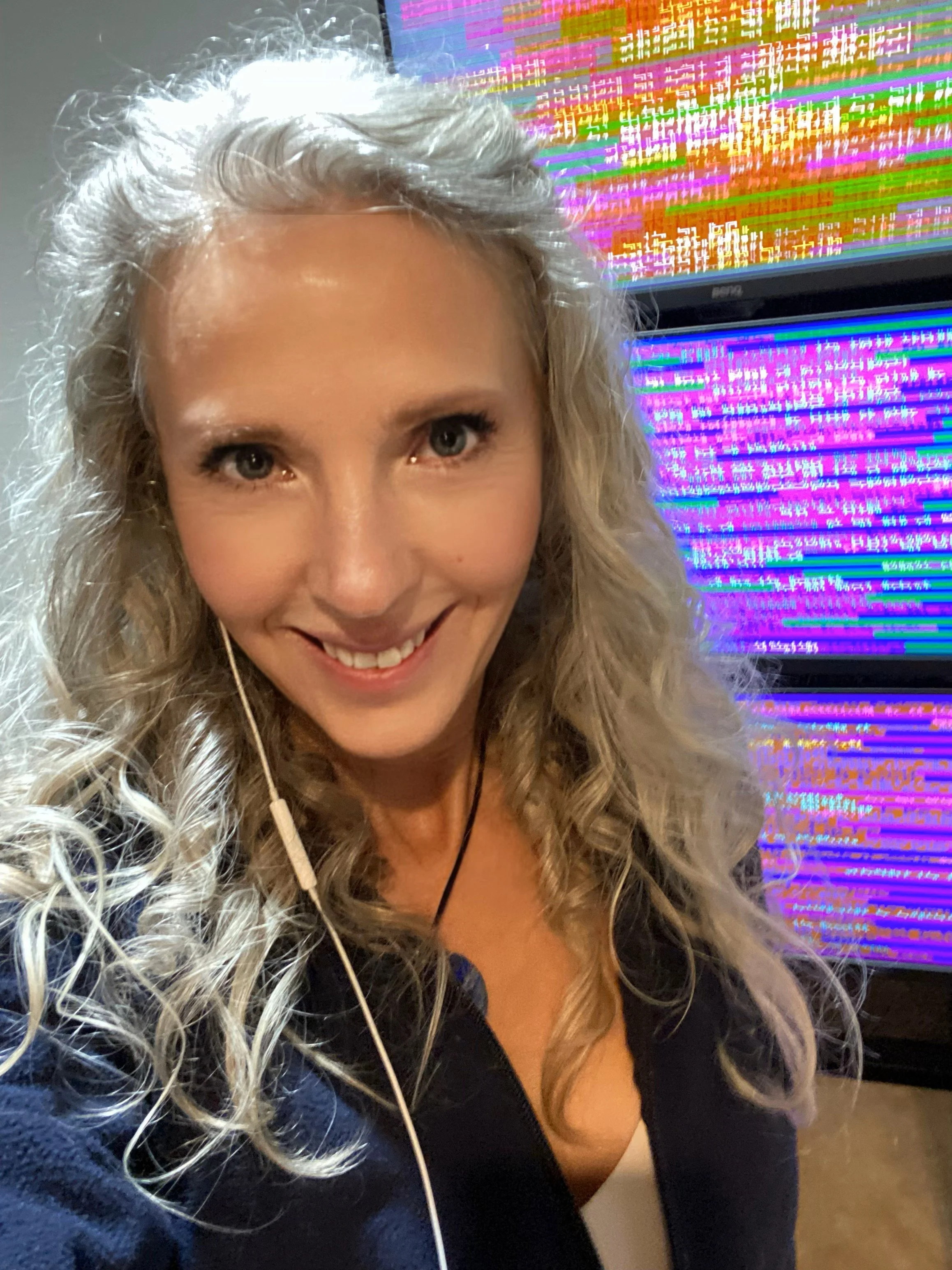 A person with curly blonde hair takes a selfie in front of The Energy Enhancement / Light System monitors displaying the colorful bio-phonic pixelated codes, graphics and geometry.