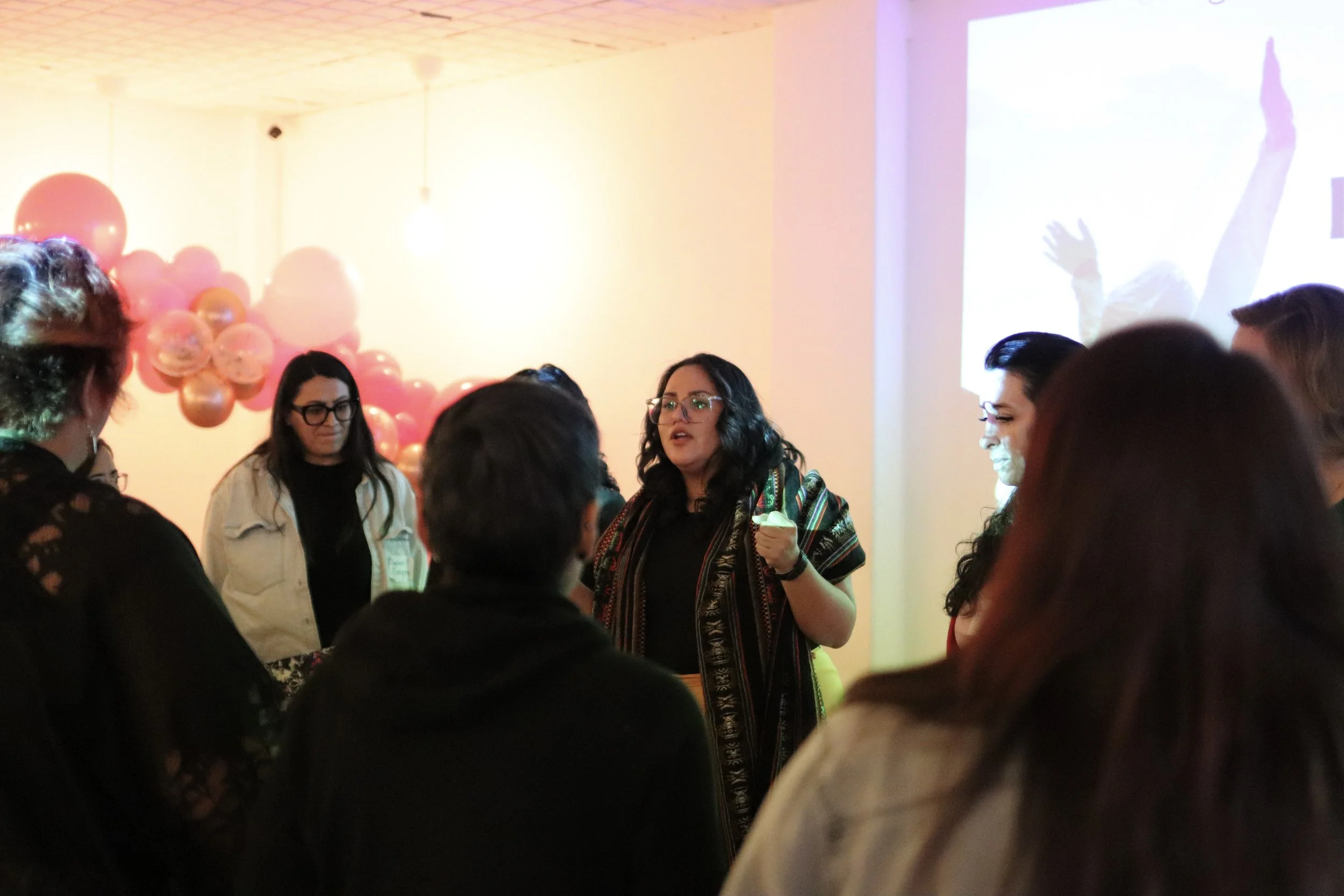 A woman with glasses and dark hair is speaking to a group of people in a room decorated with pink and peach balloons, with a large projected image or video on the wall.
