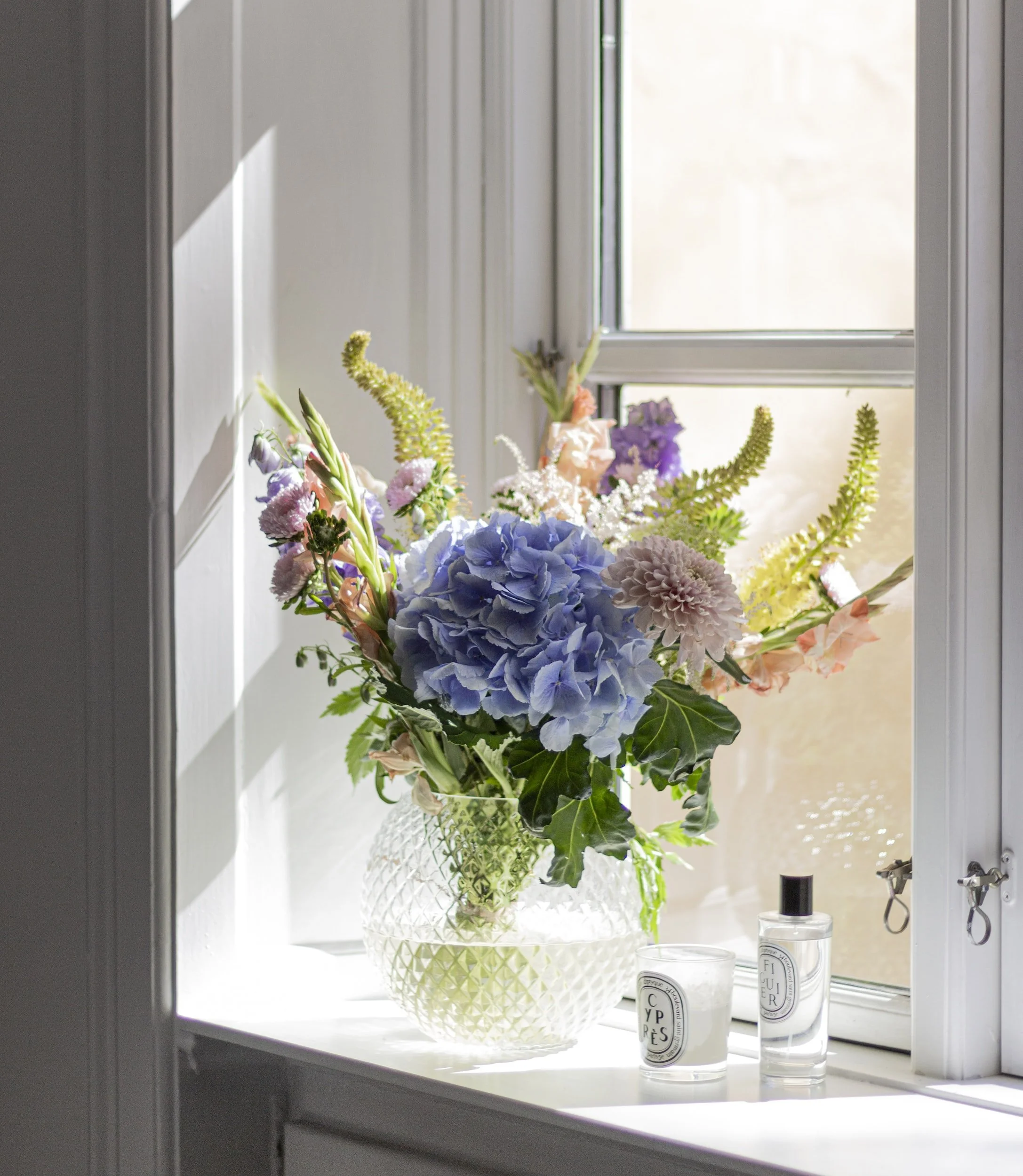 Bouquet of flowers in glass vase on windowsill with candles and diffuser