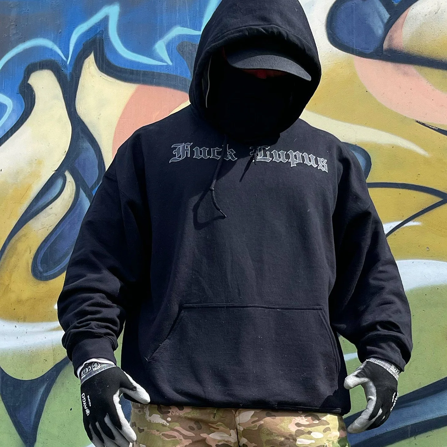 A person wearing a black hoodie with the text 'Huck Lupus' on it, black mask, gloves, and camouflage pants, standing in front of a colorful graffiti wall.