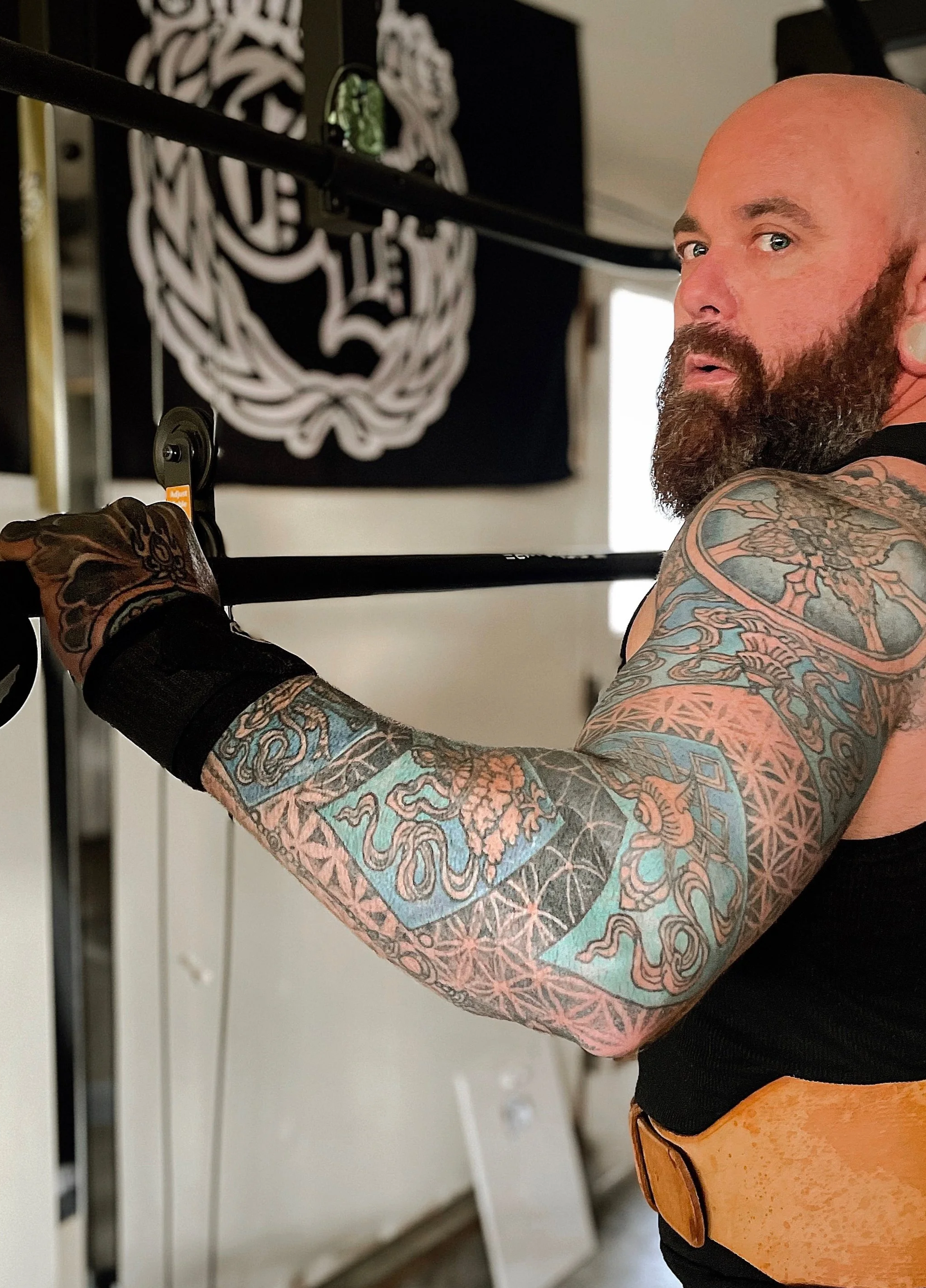 A bearded man with tattoos on his arm, wearing a black sleeveless shirt and wrist brace, appears to be pulling a bowstring, possibly preparing to shoot an arrow in a home gym.