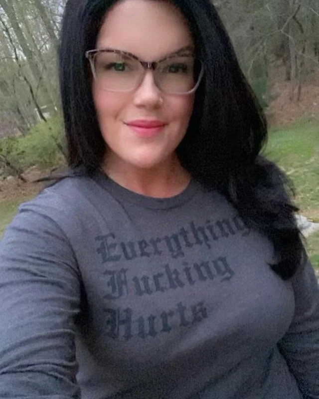 A woman with black hair wearing glasses and a dark gray T-shirt with the words "Everything Fucking Hurts" printed on it, outdoors with trees and greenery in the background.