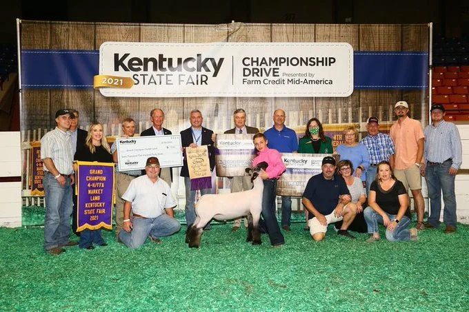 Group of people at Kentucky State Fair, celebrating victory with a sheep, awards, and a large check, in a barn-like setting with a banner overhead.