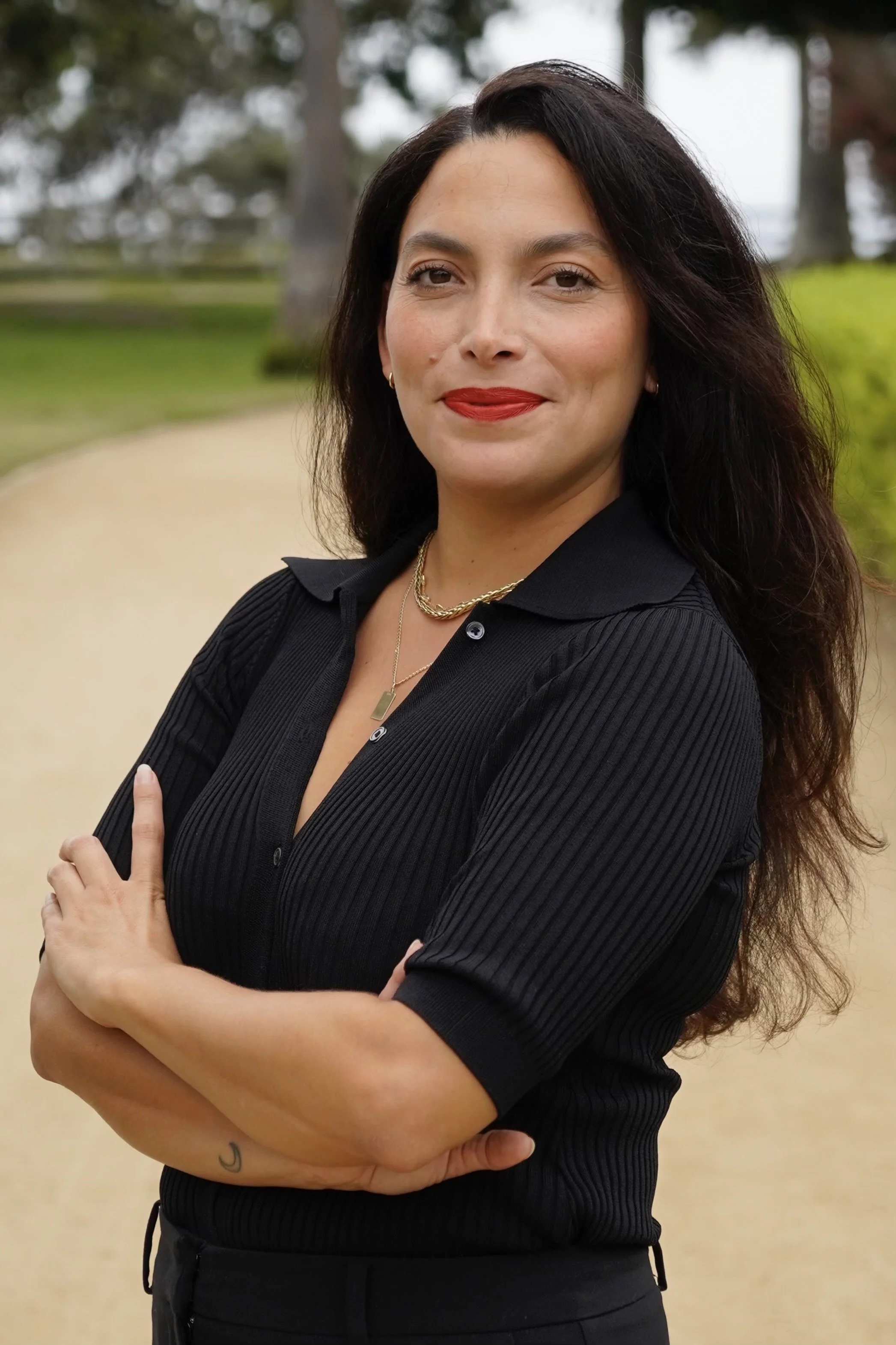 A confident woman with dark wavy hair standing outdoors with arms crossed, wearing a black ribbed shirt, red lipstick, and gold jewelry, with trees and a pathway in the background.