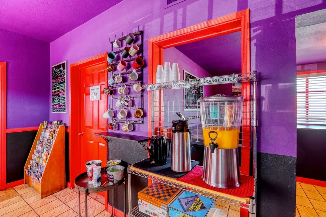Colorful room with purple walls featuring a refreshment area with mugs, coffee pots, and an orange juice dispenser. A menu board and brochure stand are also visible.