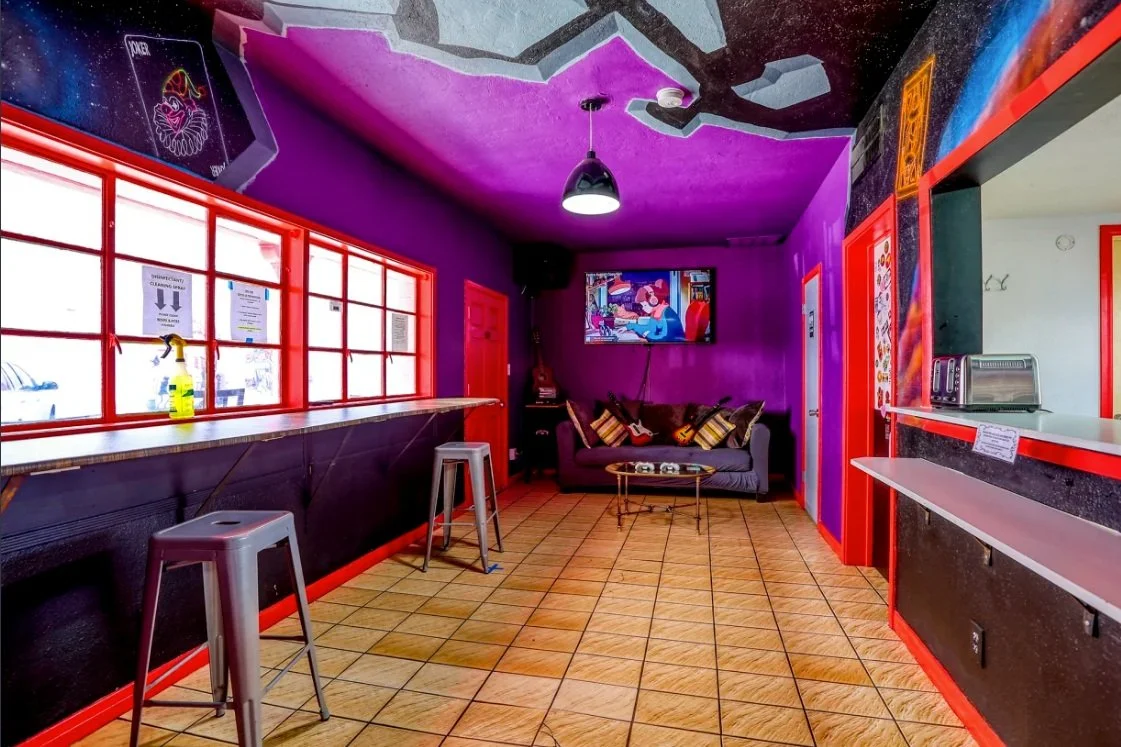 Colorful room with purple walls, red window frames, a bar counter with stools, a couch with decorative pillows, a wall-mounted TV, and tiled floor.