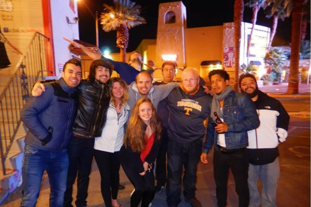 Group of nine people posing together outdoors at night, with some wearing jackets and holding drinks, in an urban setting with palm trees and lit buildings in the background.
