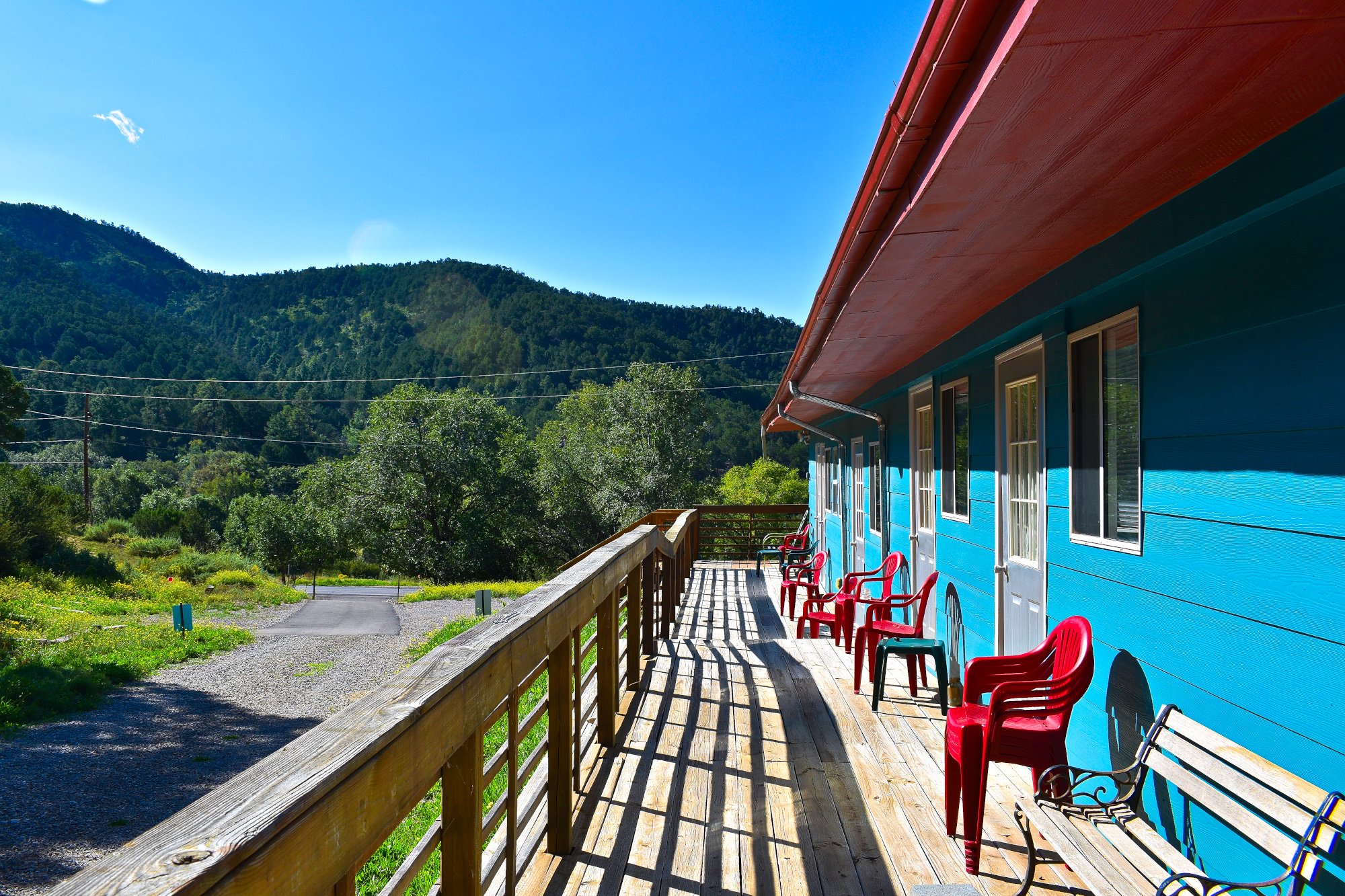 A wooden balcony with red and green chairs next to a bright blue house, overlooking a lush green landscape with hills and trees under a blue sky.