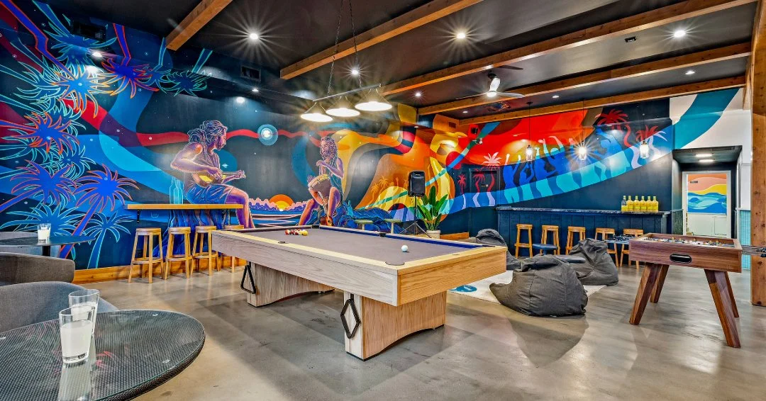 Indoor recreational room with a colorful mural, pool table, foosball table, bean bag chairs, bar stools, and tropical-themed decorations.